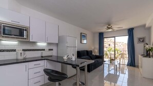 A modern kitchen with a microwave, toaster, and coffee maker, a dining area with a black sofa and table, and a balcony with a view of buildings.