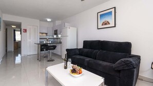 A modern living room with a black sofa, a white coffee table with fruit and a bottle, a kitchen in the background, and a framed picture on the wall.