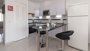 A modern kitchen with white cabinets, a stainless steel countertop, a microwave, an oven, a refrigerator, and two bar stools.