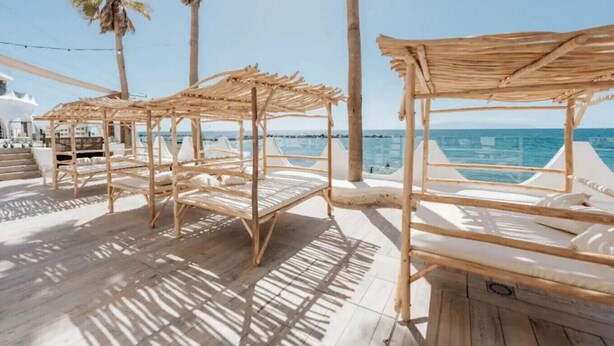 Terraza de madera con sombrillas de paja y sillas blancas acolchadas con vista a la playa.
