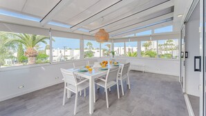 Un comedor amplio con una mesa blanca dispuesta para cuatro personas, ventanas grandes que ofrecen vistas a palmeras y edificios, y una lámpara colgante.