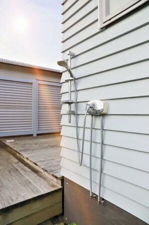 Une façade extérieure d’habitation avec une douche extérieure en métal fixée au revêtement, une terrasse en bois et une porte de garage fermée.