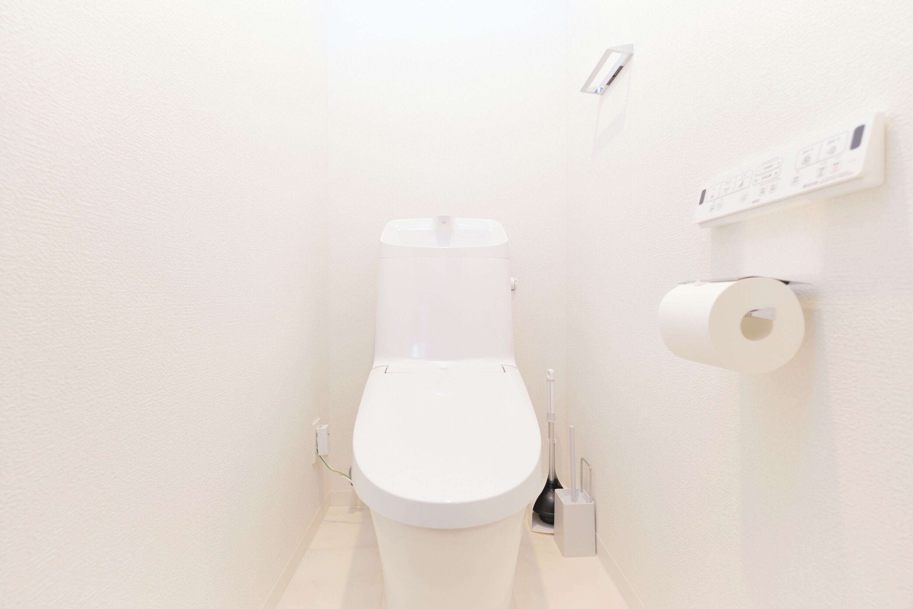 Un inodoro blanco con palanca de descarga, un dispensador de papel higiénico y un panel de control de descarga en la pared.