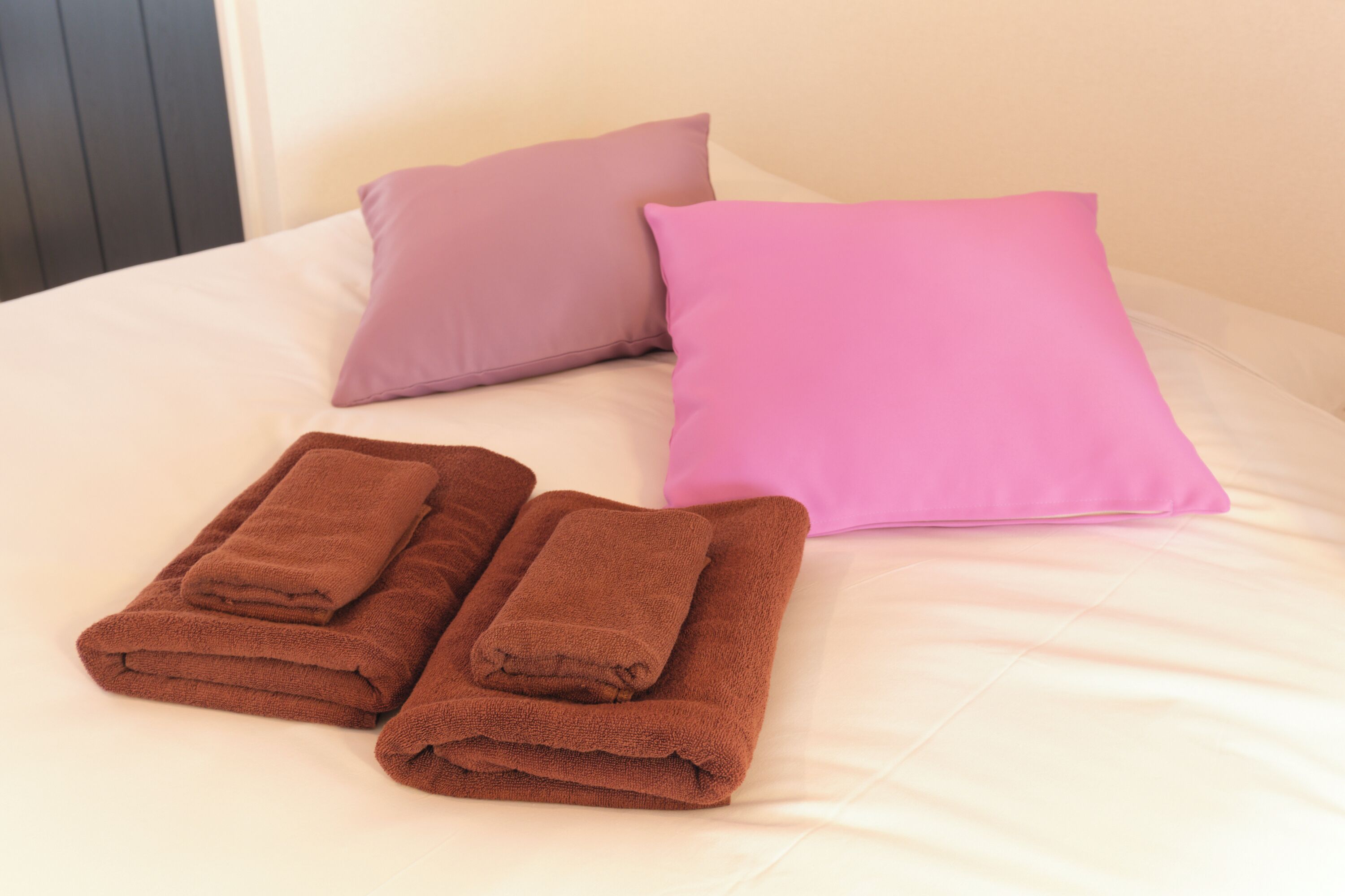 Una cama con sábanas blancas, dos almohadas (una morada y otra rosa), y dos toallas marrones dobladas.