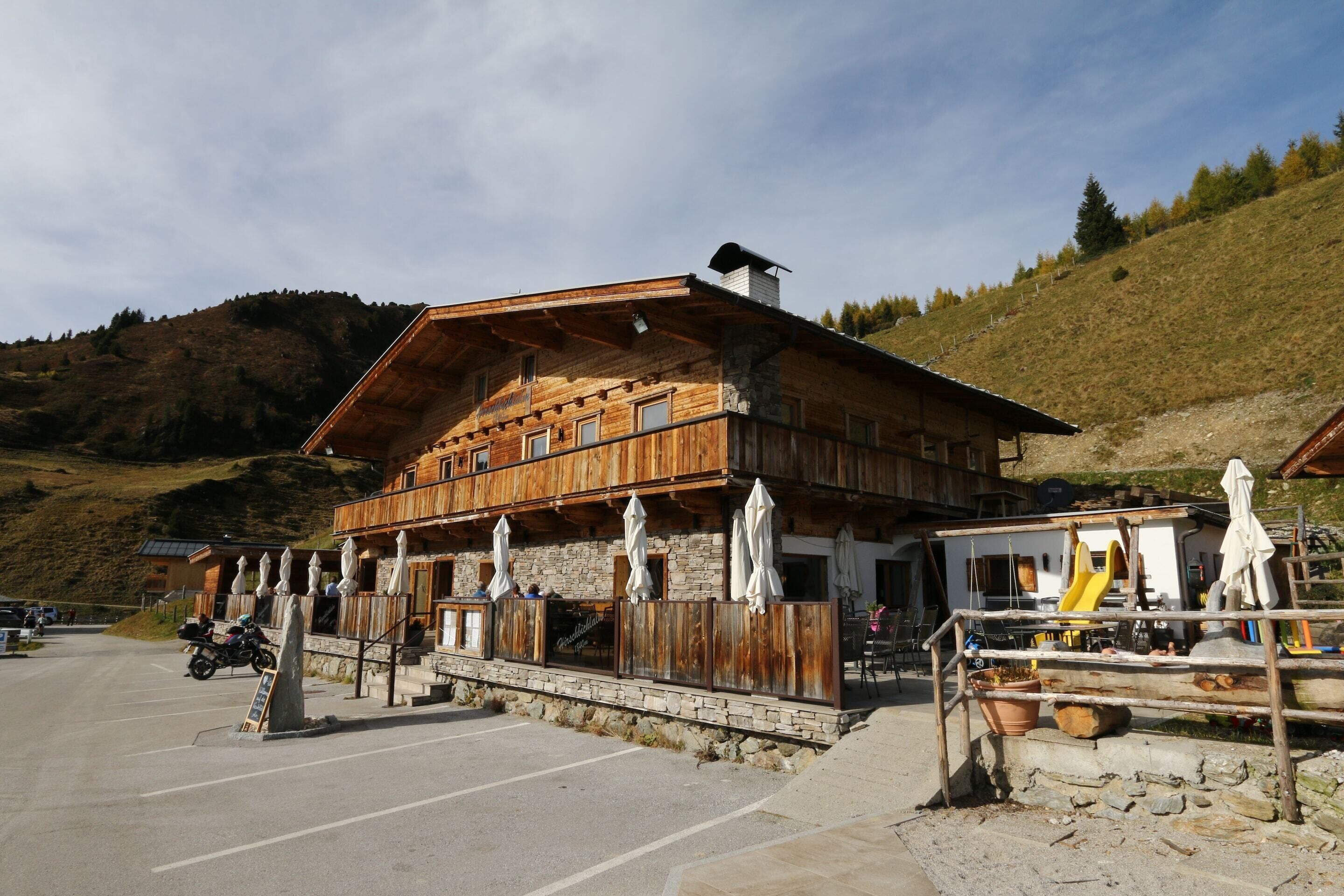 A wooden chalet with a stone foundation, outdoor seating, and a mountain backdrop.