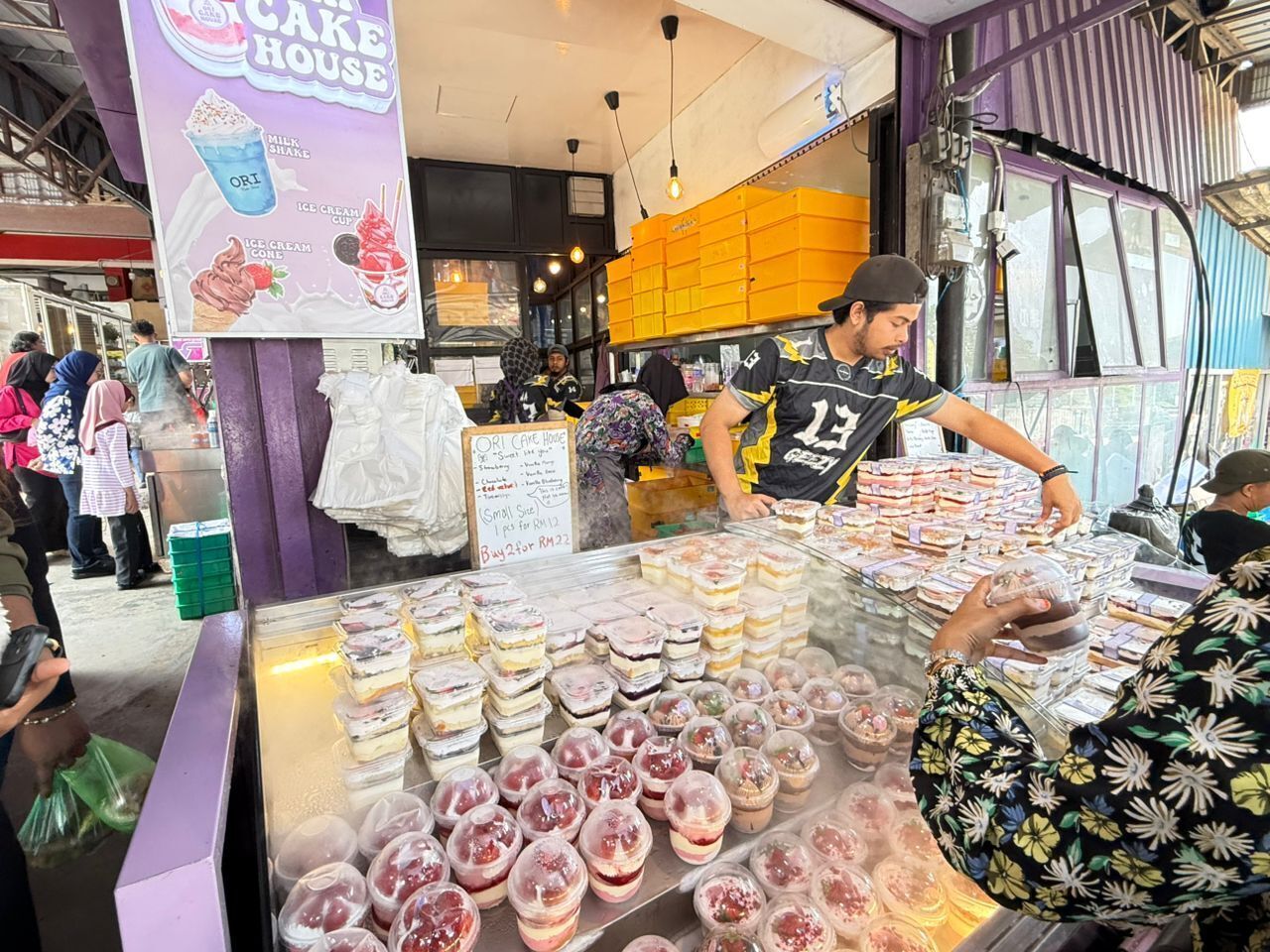 A dessert stall with various ice cream and cake options, a signboard with prices, and a busy street scene in the background.