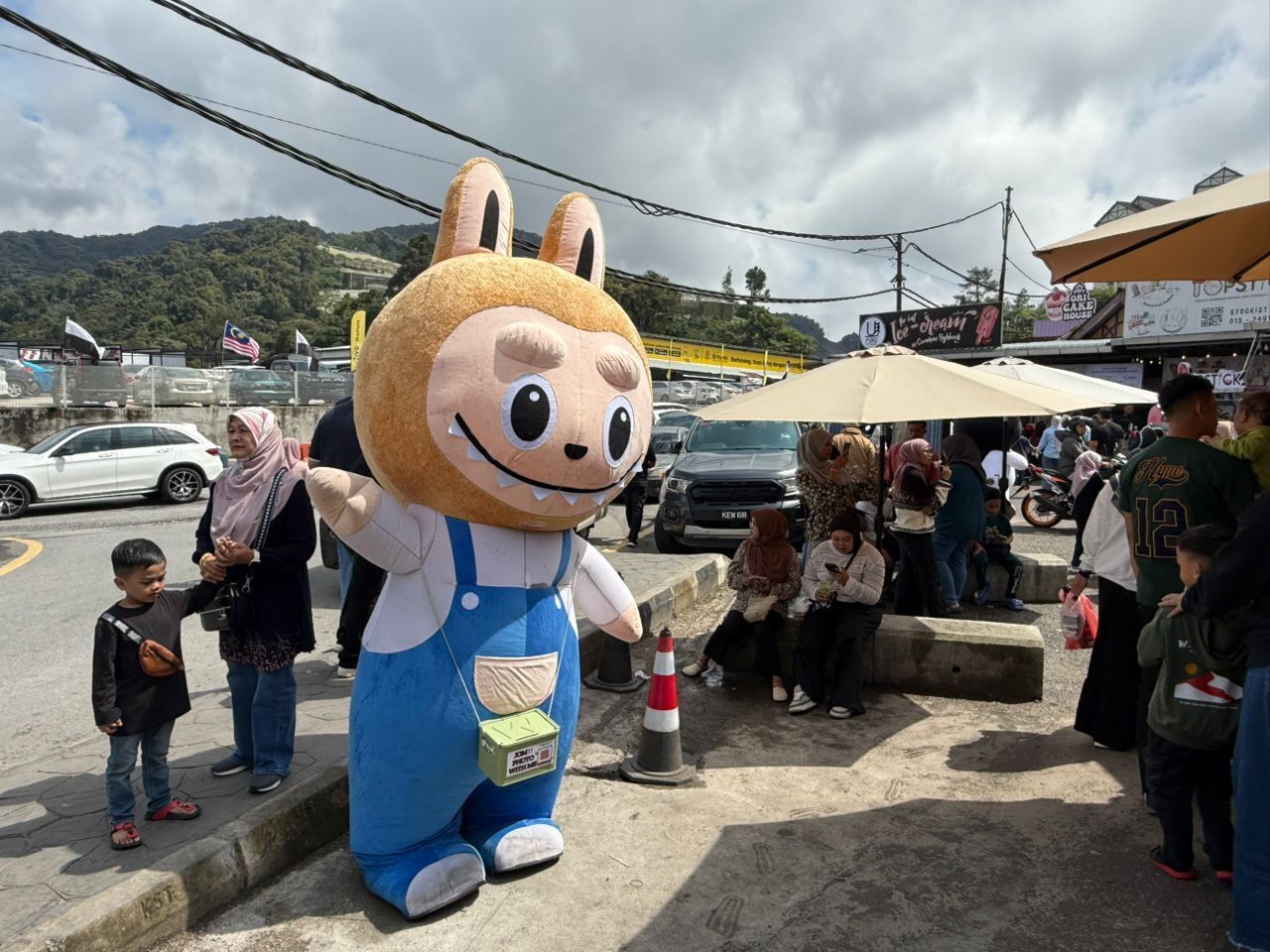 A mascot character standing on steps with a crowd of people in the background.
