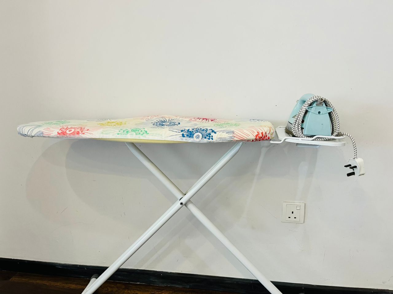 Una tabla de planchar con funda estampada y un planchador azul sobre una pared blanca.