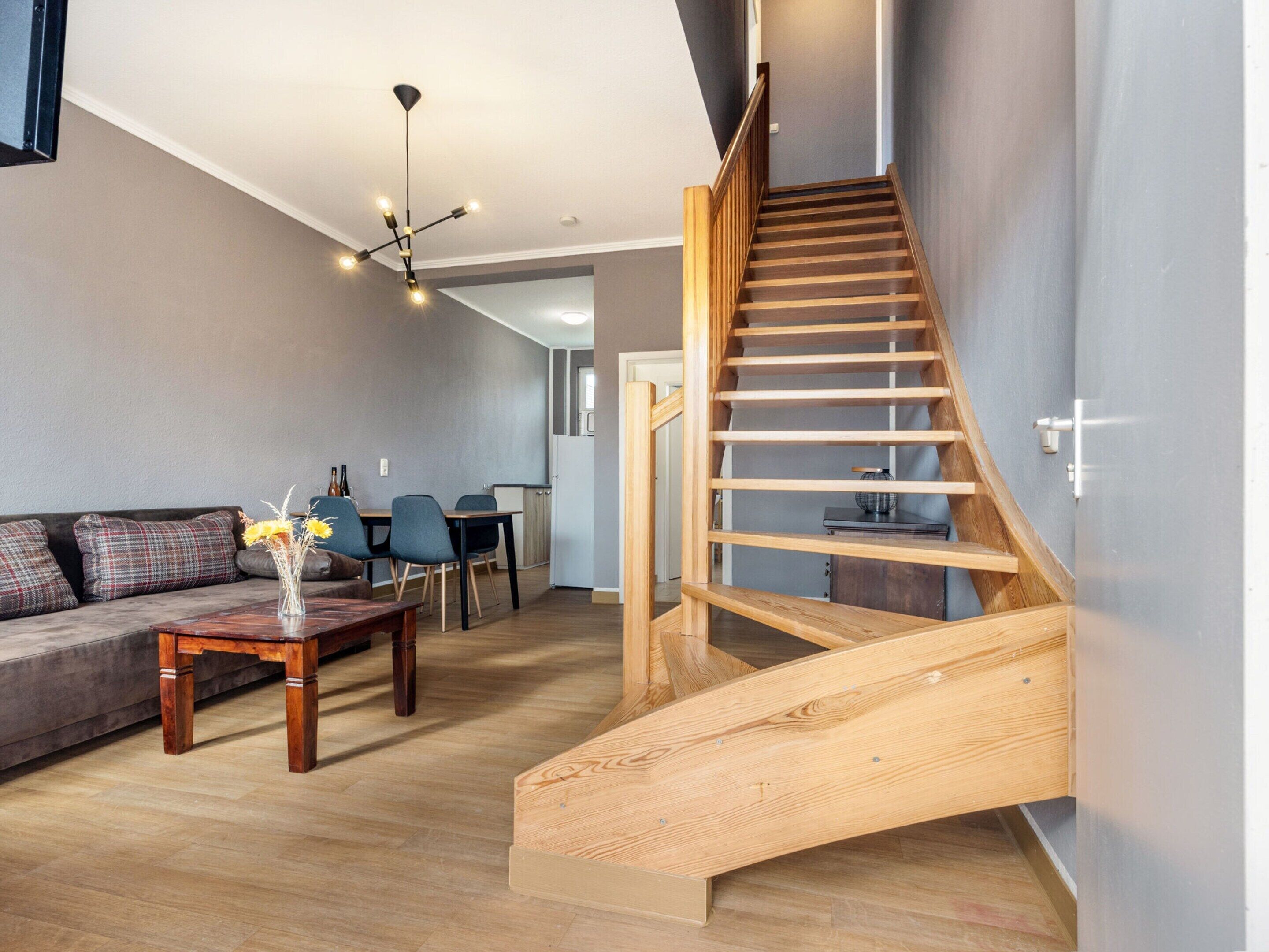 A modern living room with a wooden staircase, a dining area, and a kitchen in the background.