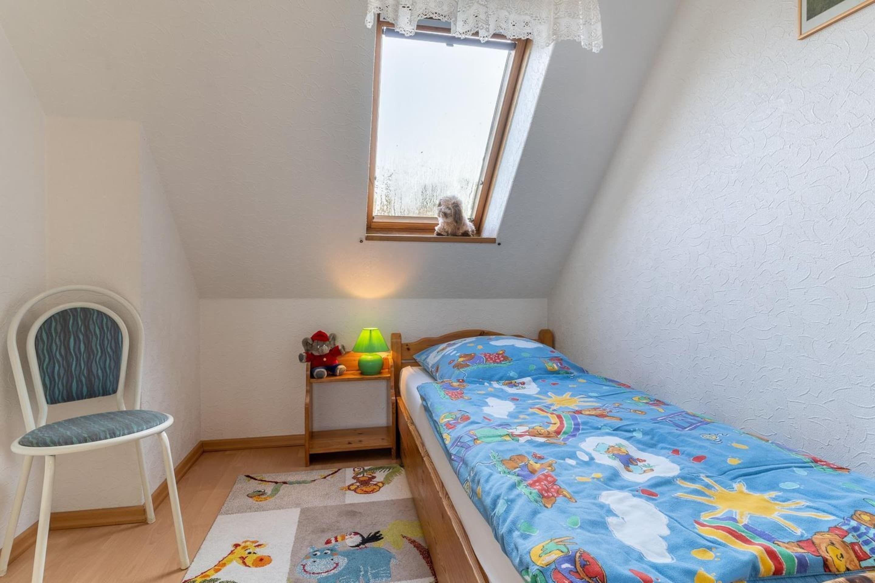 A child's bedroom with a bed, a chair, a small table with toys, a window, and a rug with cartoon characters.