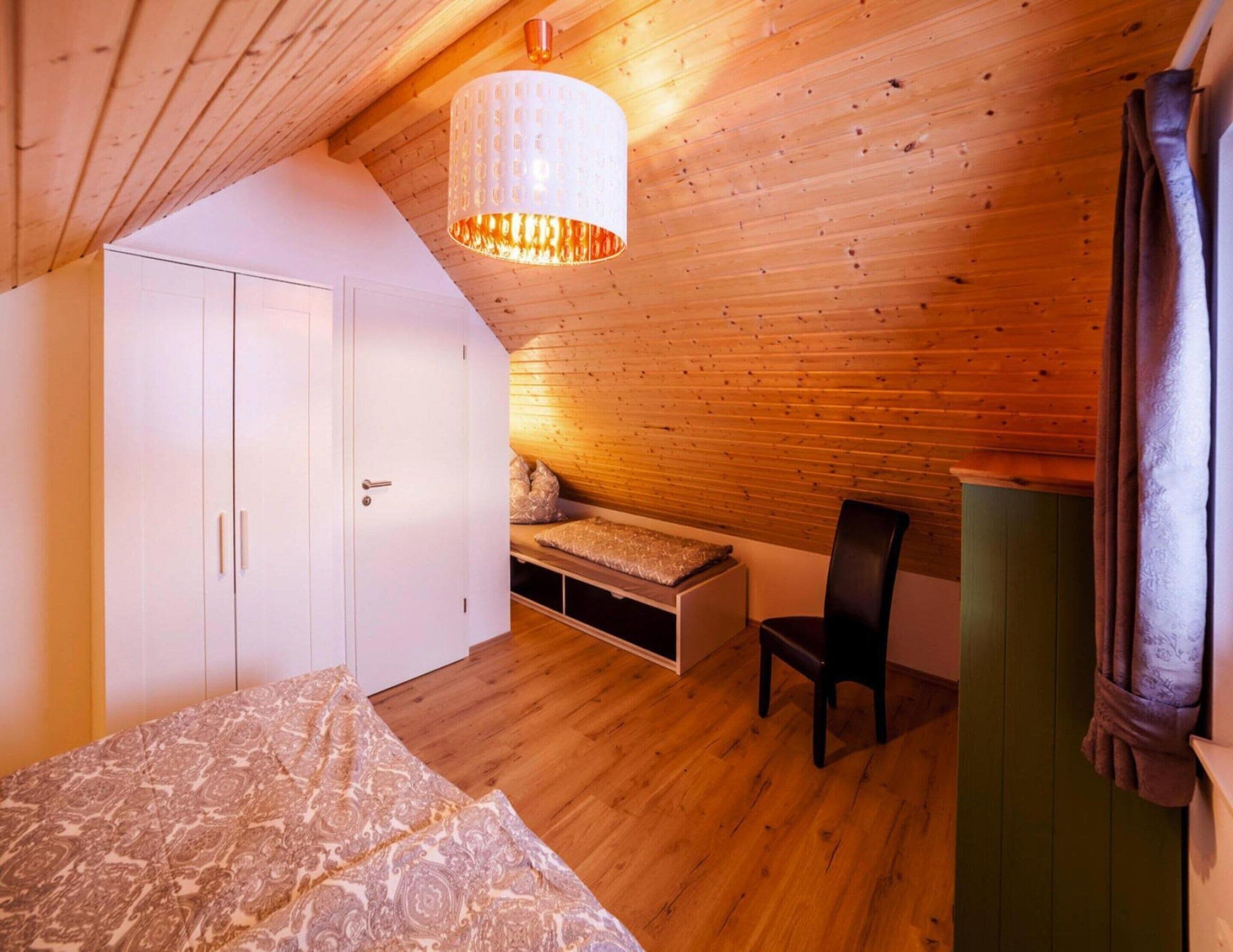 Un dormitorio con techo de madera, una cama con ropa de cama estampada, un armario blanco, un banco con cojines y una silla.