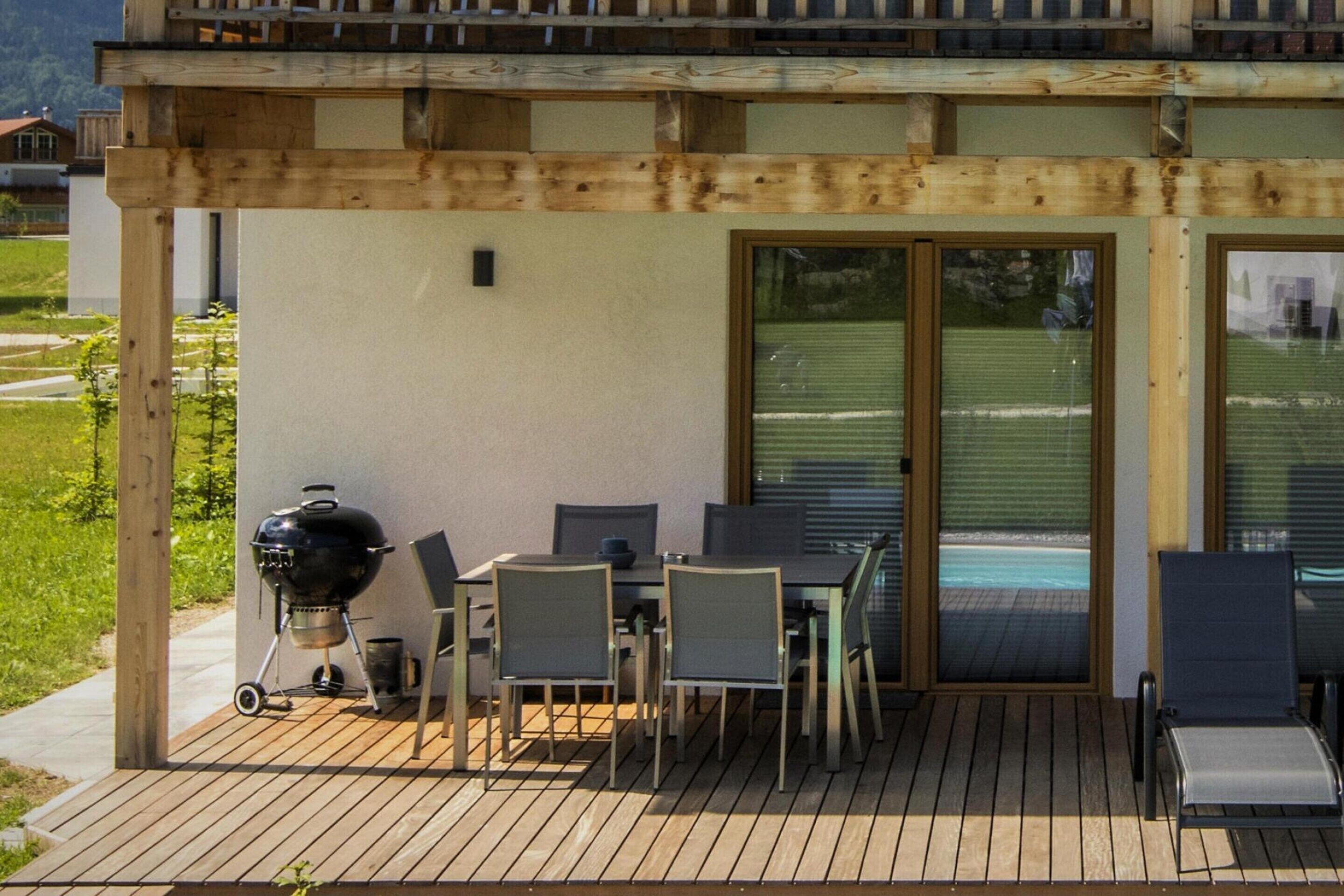 Eine überdachte Terrasse im Freien mit Tisch und Stühlen, einem Grill und Blick auf einen Swimmingpool.