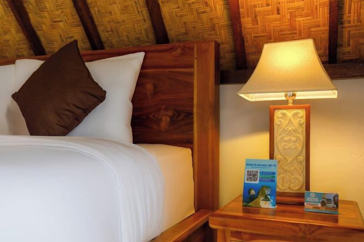 Ein Hotelzimmer mit einem hölzernen Kopfteil, einem weißen Bett mit einem braunen Kopfkissen, einem Nachttisch mit einer Lampe und Informationsbroschüren.