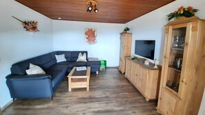 Een woonkamer met een donkerblauwe bank, een houten salontafel, een houten kast met glazen deuren, en een flatscreen televisie aan de muur.