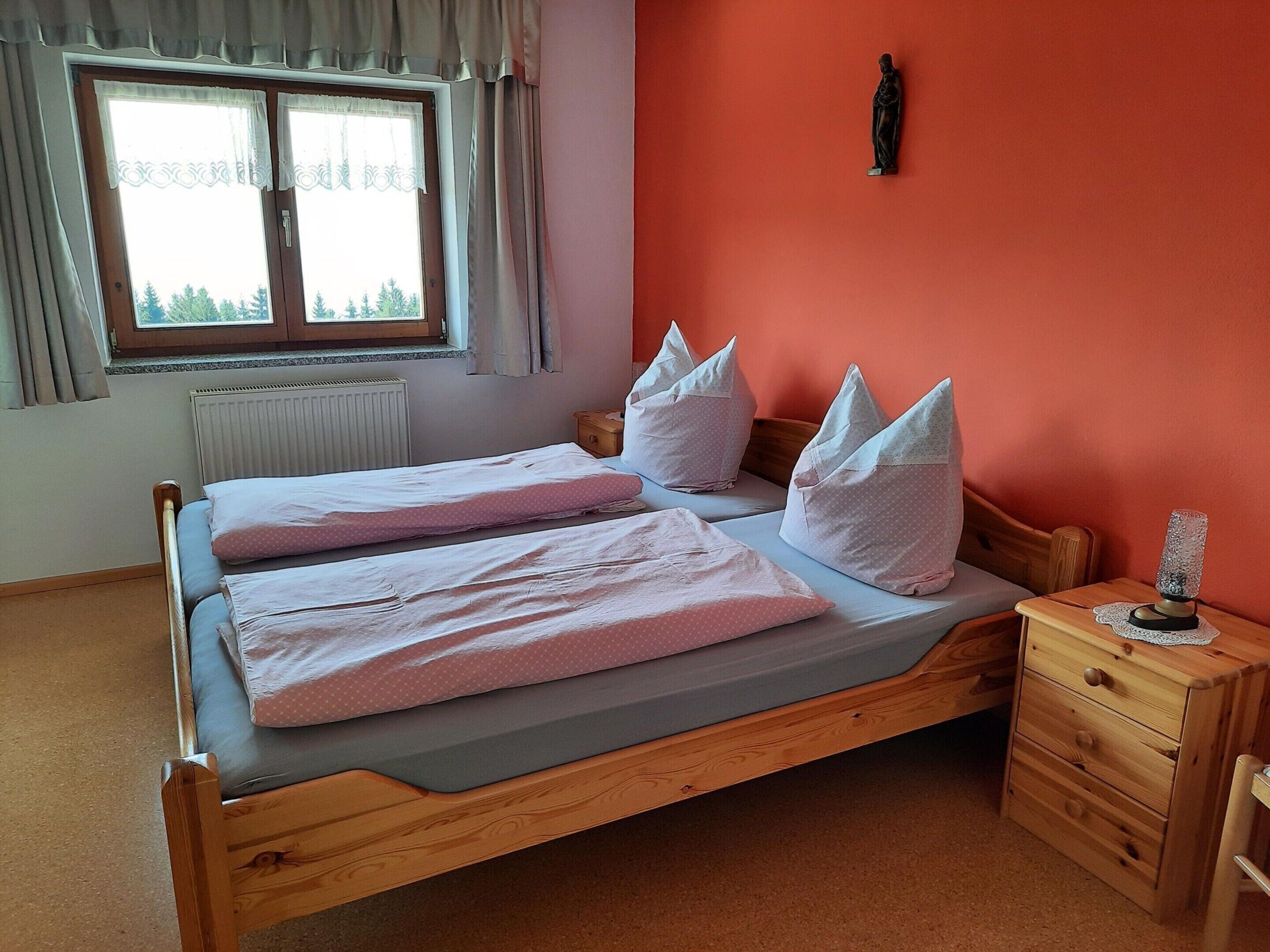 Una cama doble con dos almohadas, una mesita de noche de madera con cajones, un radiador y una ventana con cortinas.