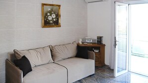 Un salon avec un canapé beige, une petite table d’appoint en bois, un tableau encadré accroché au mur et une porte-fenêtre coulissante donnant sur un espace extérieur.