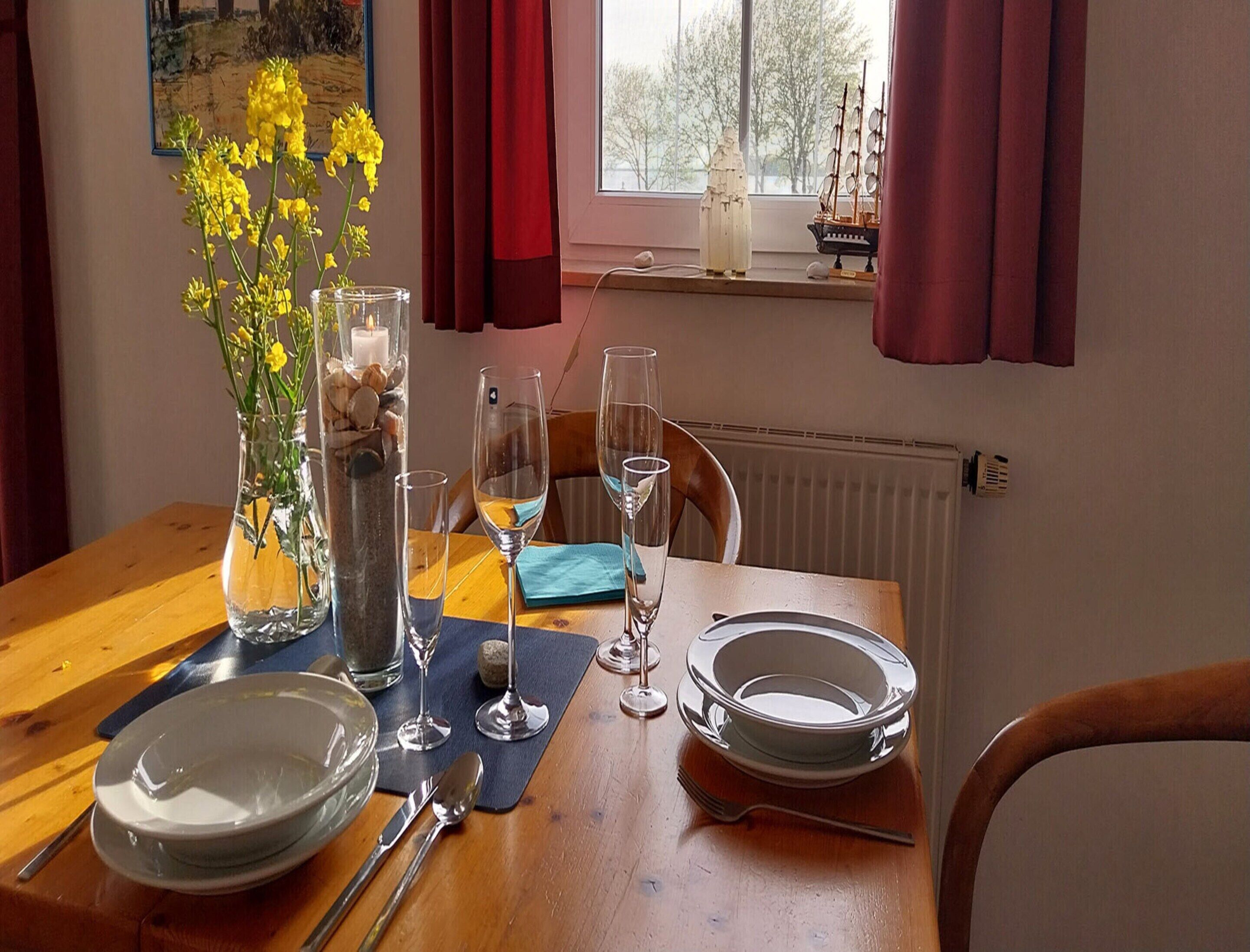 Tavola imbandita con piatti, posate e bicchieri da vino, con un vaso di fiori gialli e una candela.