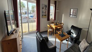 Een woonkamer met een bank, een salontafel, een televisie op een houten standaard, een eettafel met stoelen en uitzicht op een jachthaven door een schuifdeur.