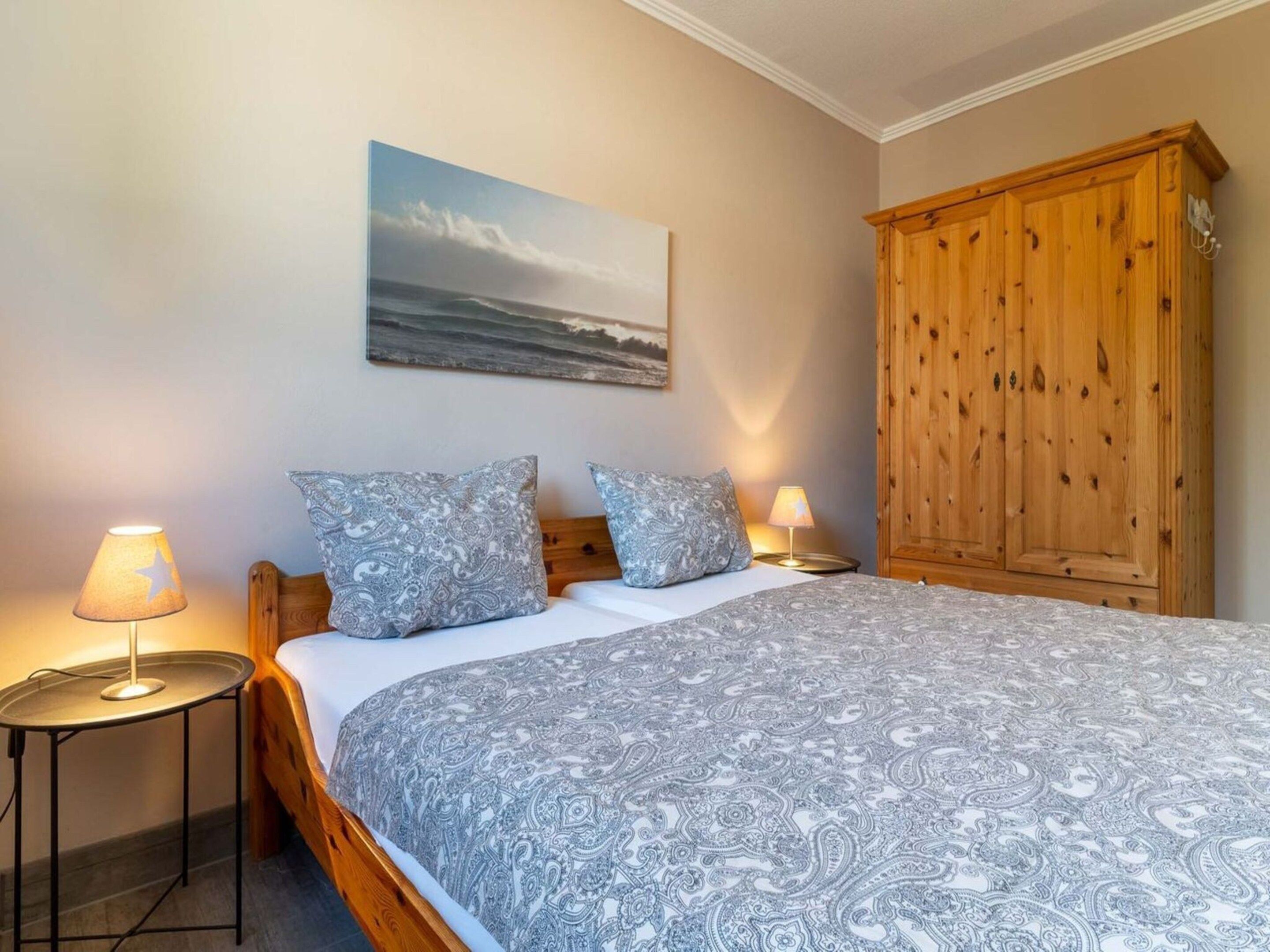 A bedroom with a bed, two bedside lamps, a wooden wardrobe, and a painting on the wall.