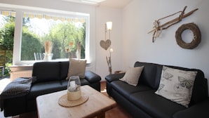 A living room with two black sofas, a wooden coffee table, and a large window with a view of greenery. - Ferienwohnung in St. Peter-ording (St. Peter-Ording)