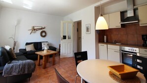 A living room with a sofa, a coffee table, and a dining table with chairs. There is a kitchen area with cabinets and a stove.