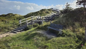 A wooden staircase leading up a hill, a bench, and a grassy area with a clear sky. - Ferienwohnung in St. Peter-ording (St. Peter-Ording)