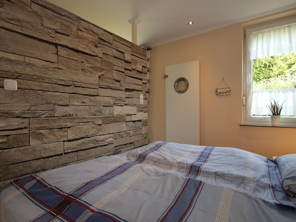 Une chambre avec un mur en pierre, un lit recouvert d’une courtepointe à rayures, une fenêtre avec une plante et une porte ornée d’un miroir rond.