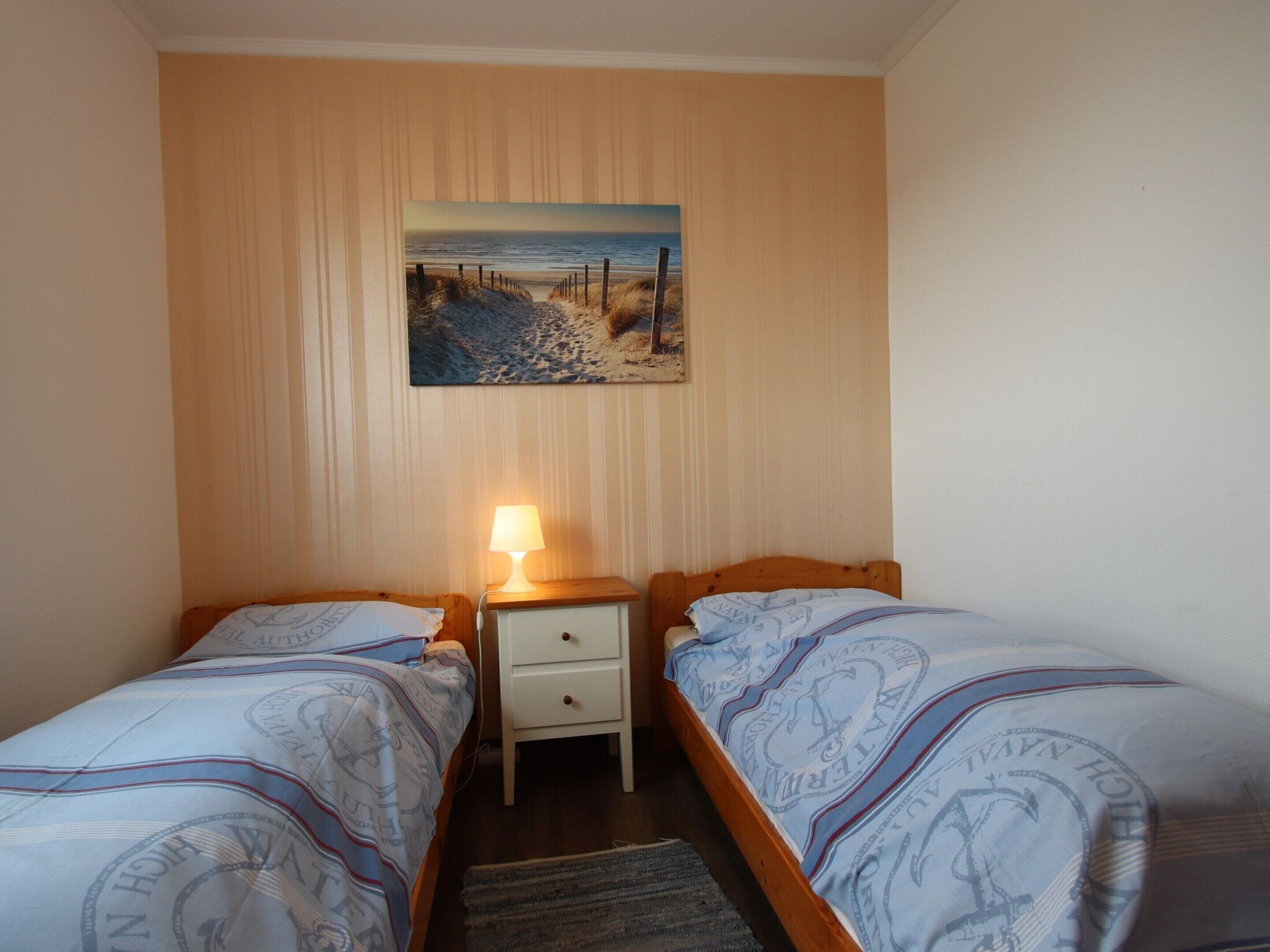 A bedroom with two single beds, a nightstand with a lamp, a painting of a beach scene, and a wooden floor.