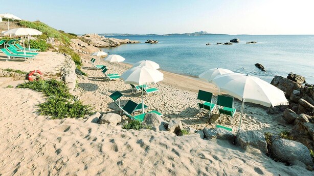 Una spiaggia con lettini verdi e ombrelloni bianchi, un litorale roccioso e un mare calmo.