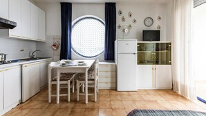 Kompakti keittiö, jossa on valkoiset kaapit, ruokapöytä tuoleineen, jääkaappi ja ikkuna, jossa on verhot.