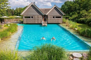 A swimming pool with two people swimming, surrounded by a house with a thatched roof and a wooden deck. - Appartement am Plauer See in Ganzlin (Ganzlin)