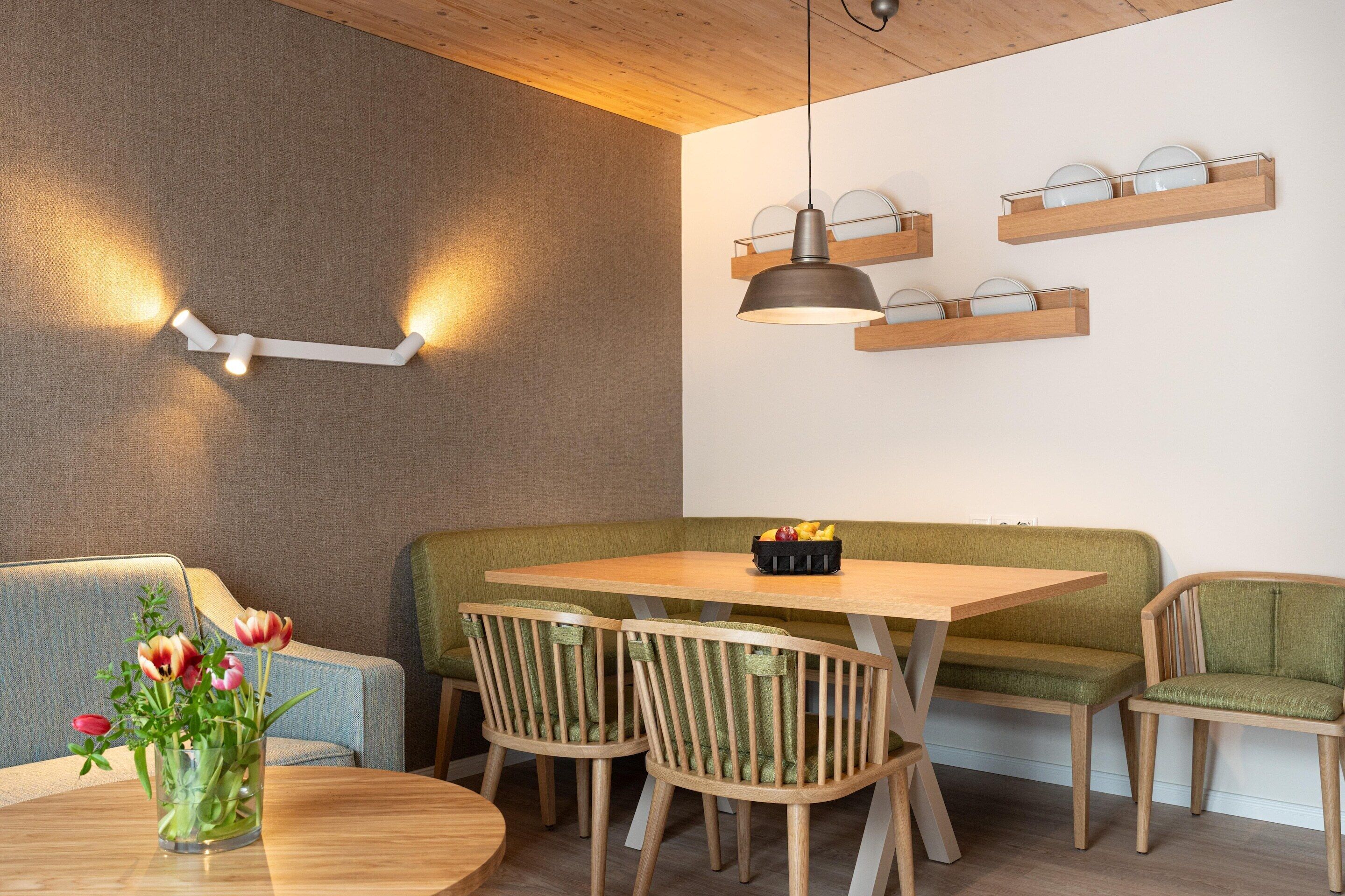 A dining area with a wooden table, green cushioned bench, and chairs. There are wall-mounted shelves with plates and a pendant light above the table.