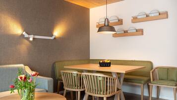 A dining area with a wooden table, green cushioned bench, and chairs. There are wall-mounted shelves with plates and a pendant light above the table.