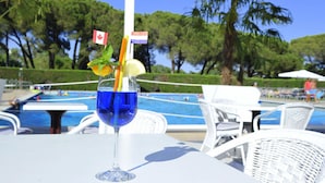 A clear glass with a blue drink, mint leaves, and a flag, placed on a white table near a pool with chairs and palm trees in the background. - New Apartment in Baia Domzia Near the sea (Sessa Aurunca)