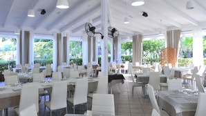 A spacious dining area with white tables and chairs, large windows, and ceiling fans. - New Apartment in Baia Domzia Near the sea (Sessa Aurunca)