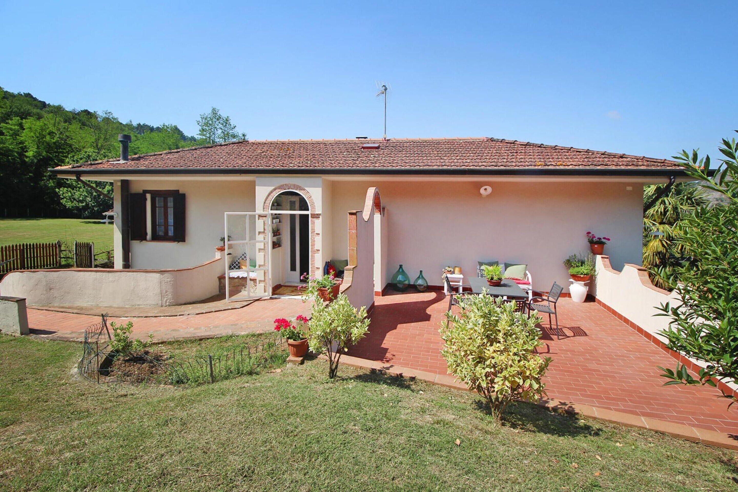 Uma casa térrea com telhado de telhas, uma porta de entrada e um pequeno alpendre com uma mesa e cadeiras.