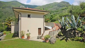 Edificio de dos pisos con balcón, rodeado de un jardín cuidado y un paisaje montañoso.