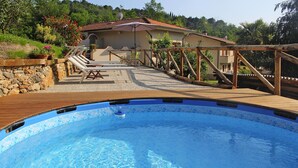 Una piscina con cubierta de madera, un muro de piedra y una casa con techo de tejas al fondo.