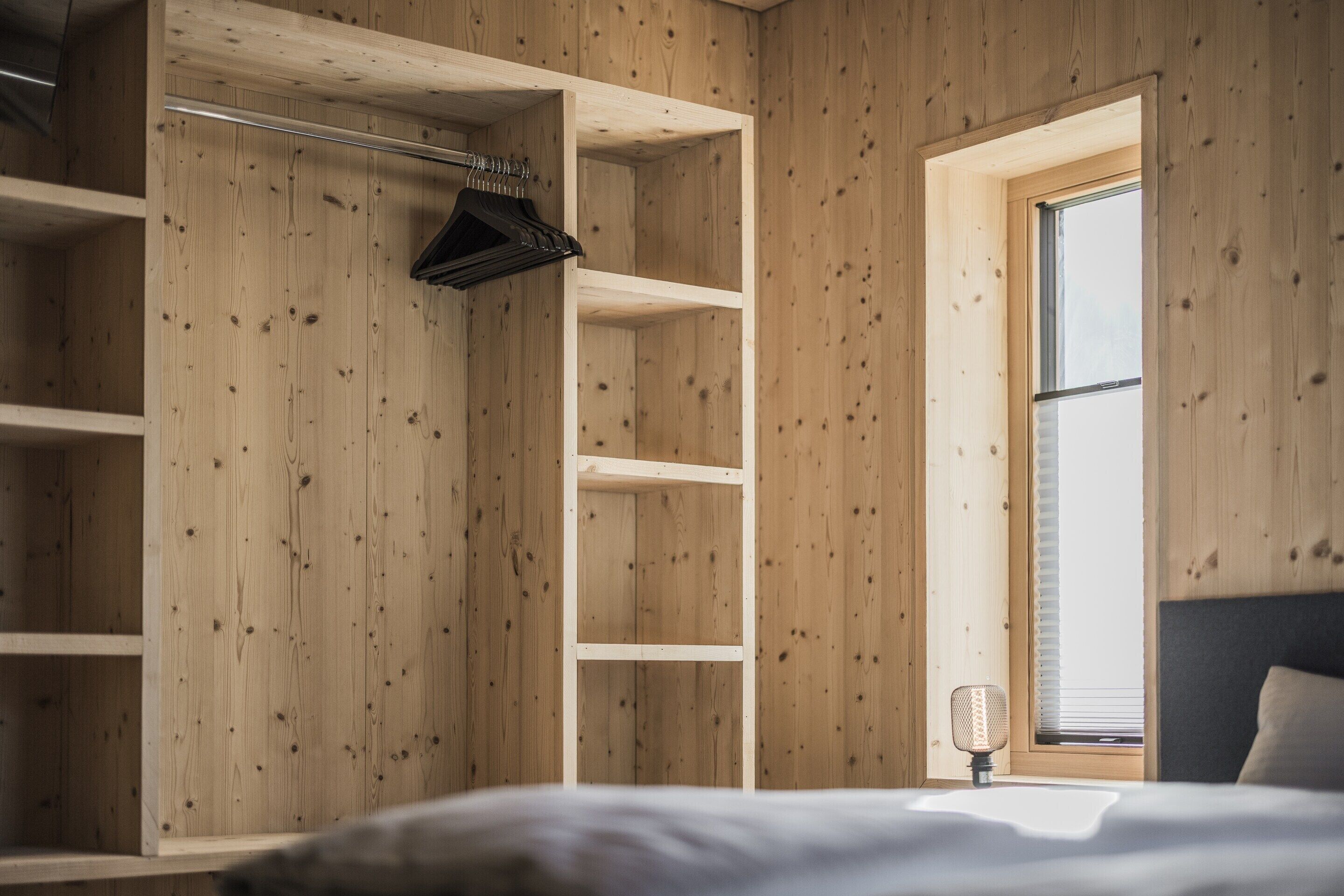 Ein Schlafzimmer mit Holzwänden, einem Bett, einem Fenster mit Jalousien und einem Kleiderständer mit Kleidung.