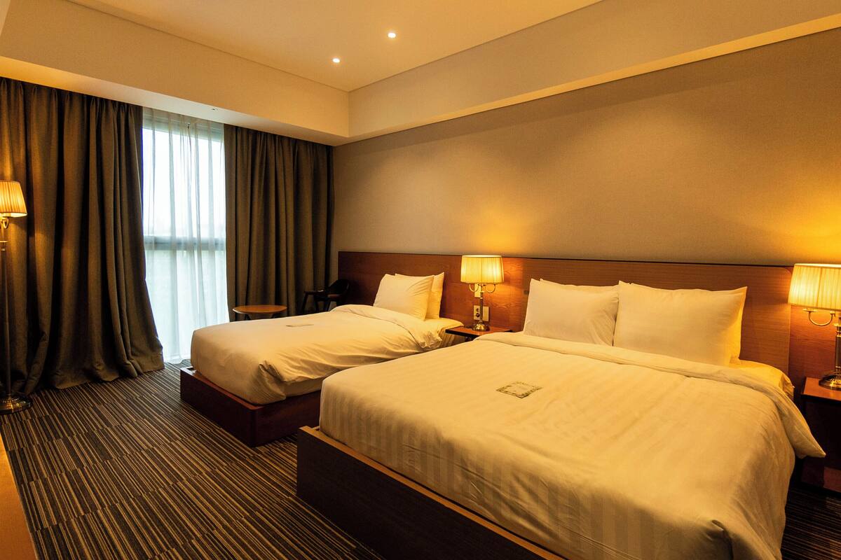 A hotel room with two beds, a large window with curtains, and two bedside lamps.