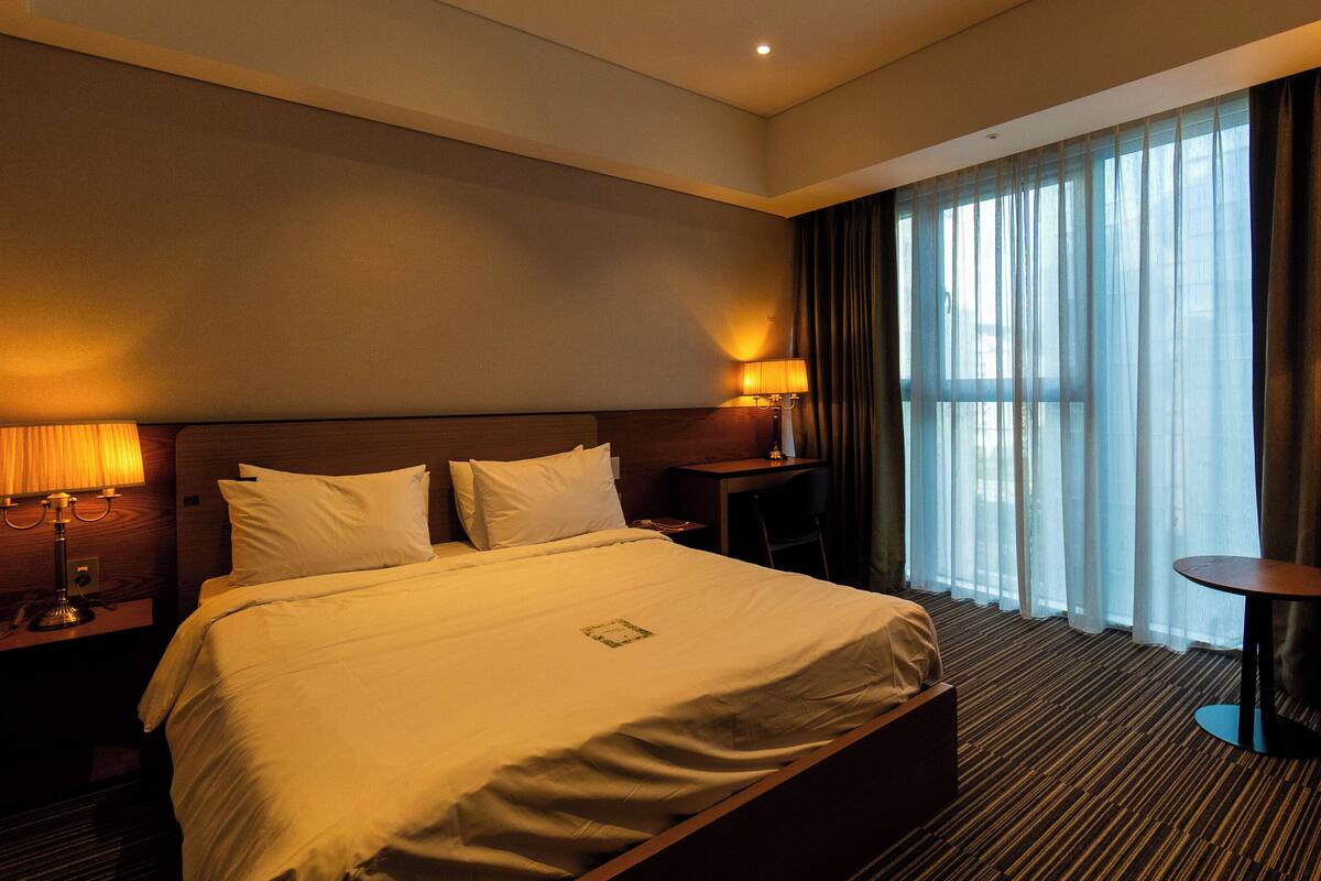 A hotel room with a large bed, two bedside tables with lamps, a desk, and a chair. There is a large window with curtains and a view of the outside.