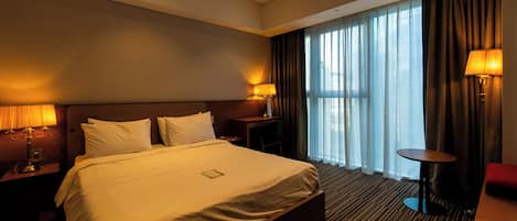 Une chambre d’hôtel avec un grand lit, deux tables de chevet avec des lampes, un bureau et une chaise. Il y a une grande fenêtre avec des rideaux et une vue sur l’extérieur.
