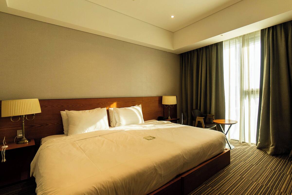 A hotel room with a large bed, two bedside tables with lamps, a small round table, a chair, and a view of the outdoors through sheer curtains.