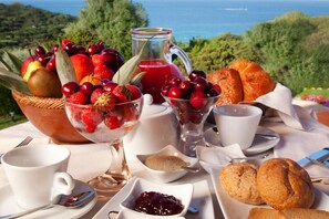 A breakfast setting with fruits, pastries, and beverages on a table with a scenic view. - Holiday Residence Punta Falcone Near Beach (Santa Teresa di Gallura)