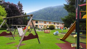 A playground with swings and slides, a building with a green roof in the background, and a mountain. - Flats Birkenhof, Dobriach Millstatter See (Radenthein)