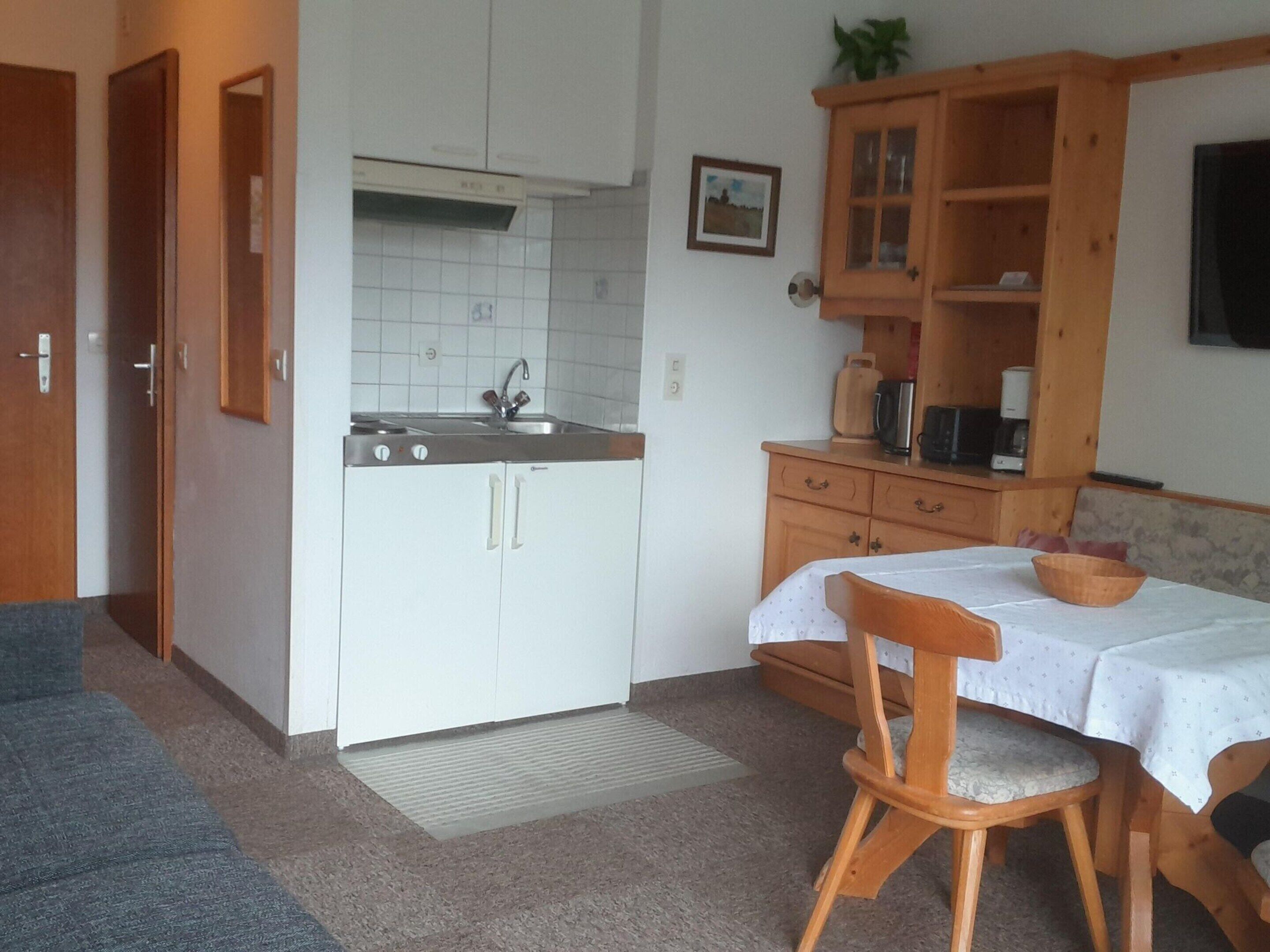 Une cuisine compacte avec des meubles blancs, un évier et un meuble en bois sur lequel est posée une plante. On y trouve également une table à manger recouverte d’une nappe blanche et une chaise en bois.