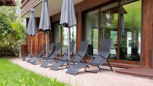 A row of sun loungers with umbrellas, a wooden fence, and a glass door leading to a garden. - Apartment in Wildschonau by Schatzberg Lift (Wildschönau)