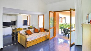 A compact living space with a kitchenette, a sofa bed with pillows, a dining area, and a view of the garden through an open door. - Apartment in Italy Near Sandy Beach (San Michele al Tagliamento)