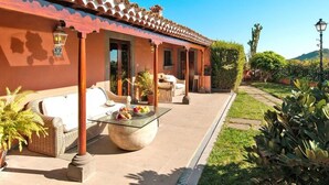 A covered outdoor seating area with wicker furniture and a glass-top table, surrounded by potted plants and a well-maintained garden. - Villa Mansion Bandama, Santa Brigida (Santa Brigida)