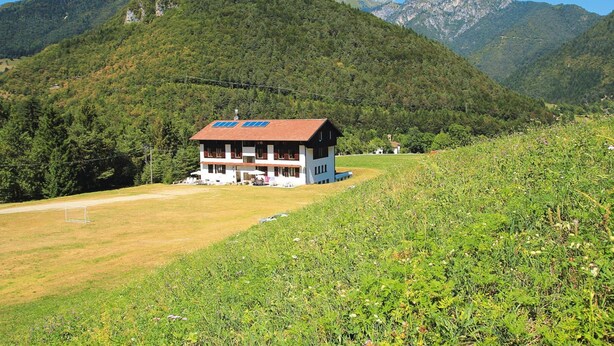 Una casa de dos pisos con techo rojo y paredes blancas, ubicada en una zona montañosa con cielo despejado.